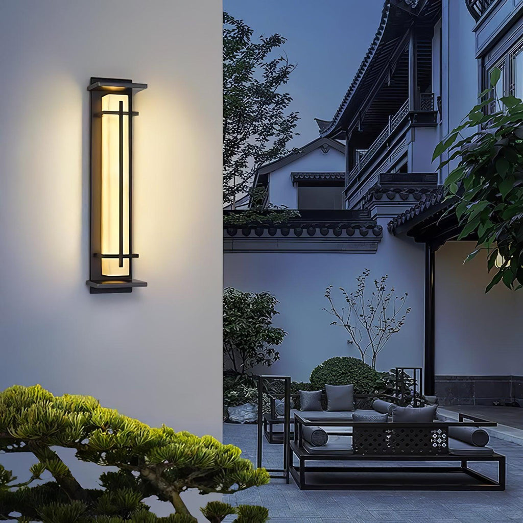 A glowing Clérachic wall light on a pale wall, with a pine tree and a courtyard seating area in the dusk behind.