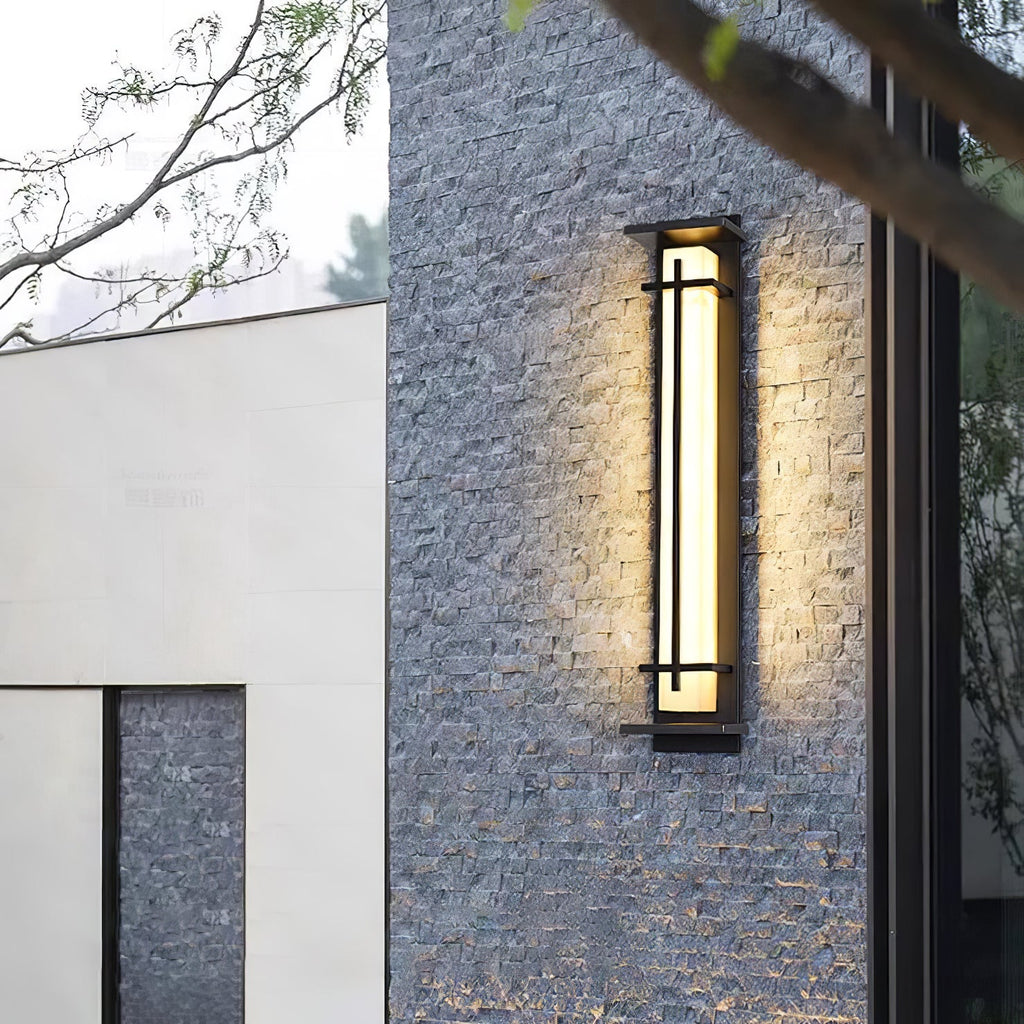 This Clérachic wall light glows against a rough grey stone wall, beside a pale rendered wall and bare branches.