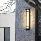 This Clérachic wall light glows against a rough grey stone wall, beside a pale rendered wall and bare branches.