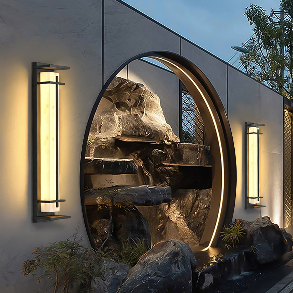 A pair of lit Clérachic wall lights flanking a round moon gate that frames a rock garden, on a long grey wall at dusk.
