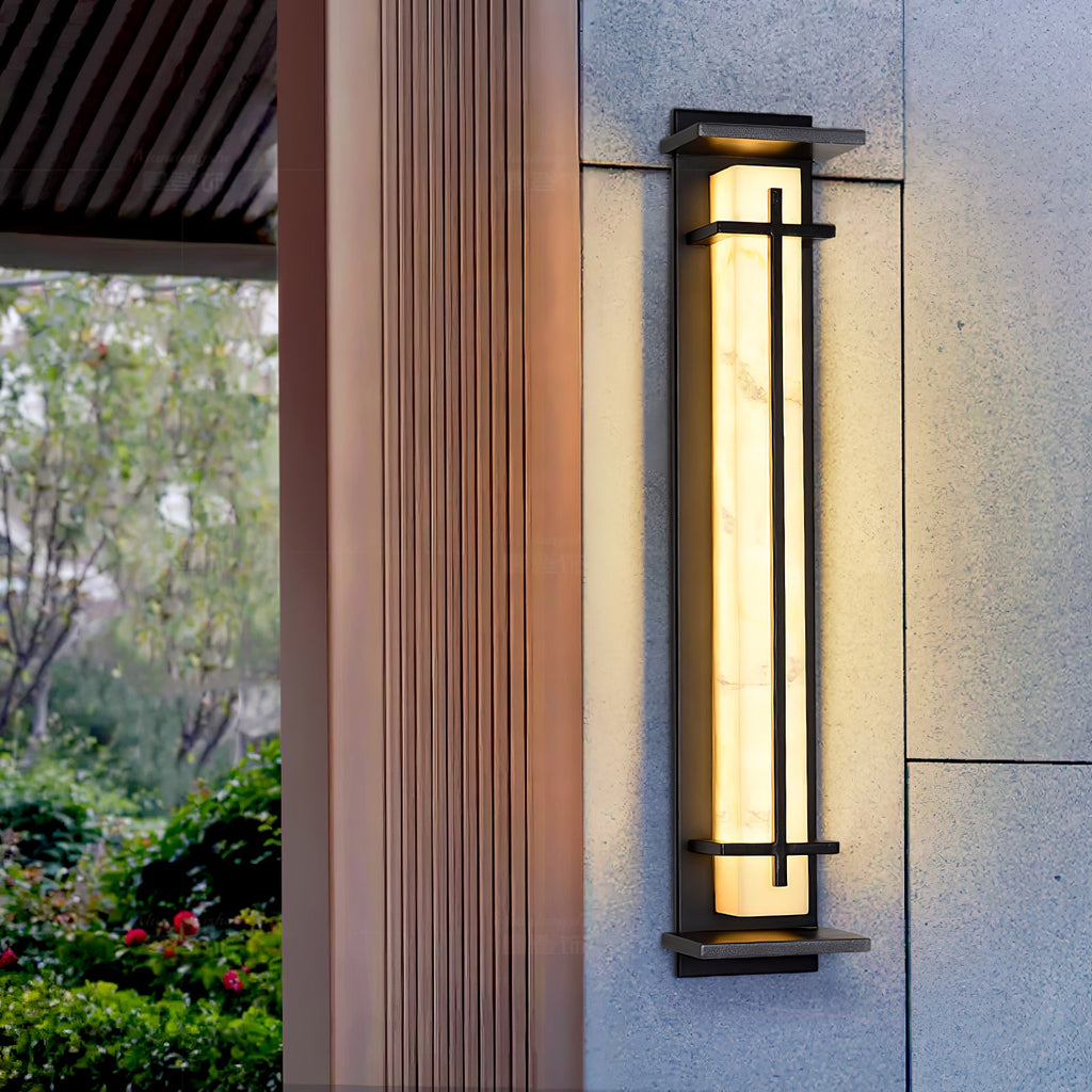 A tall Clérachic wall light glowing beside a timber-clad column, with green shrubs and a tree visible in the background.