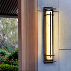 A tall Clérachic wall light glowing beside a timber-clad column, with green shrubs and a tree visible in the background.