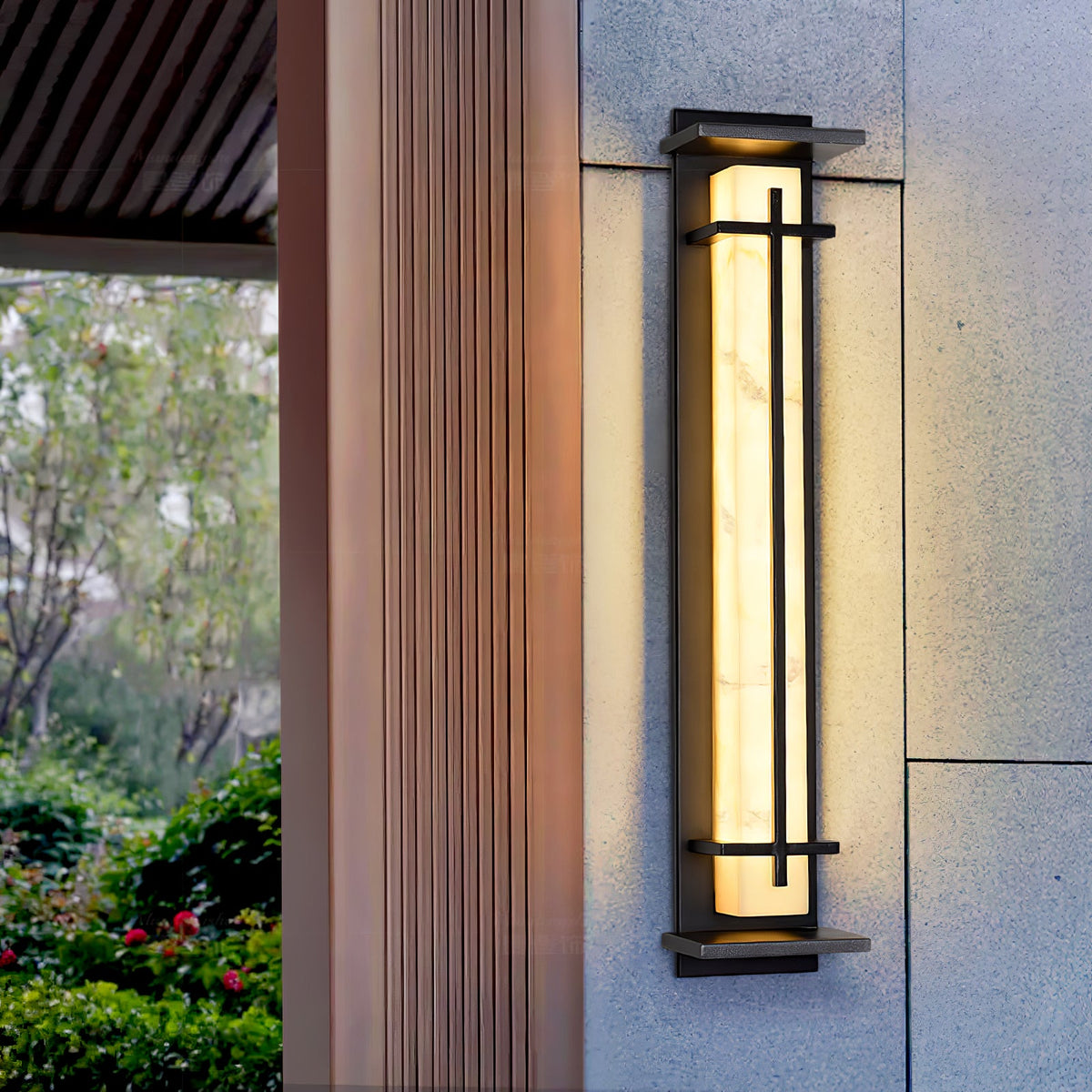 A tall Clérachic wall light glowing beside a timber-clad column, with green shrubs and a tree visible in the background.