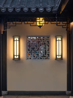 A pair of lit Clérachic wall lights framing a lattice window panel on a cream wall, under a tiled eave with a hanging lantern.