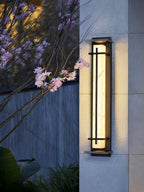 A Clérachic wall light glowing on a pale stone wall beside pink blossom branches at dusk.