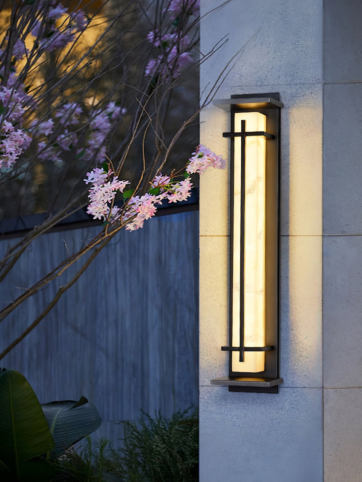 A Clérachic wall light glowing on a pale stone wall beside pink blossom branches at dusk.