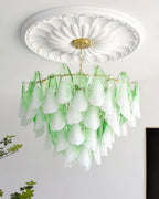 The Clérachic chandelier in sage green glass petals lit in a room corner beside a potted plant.