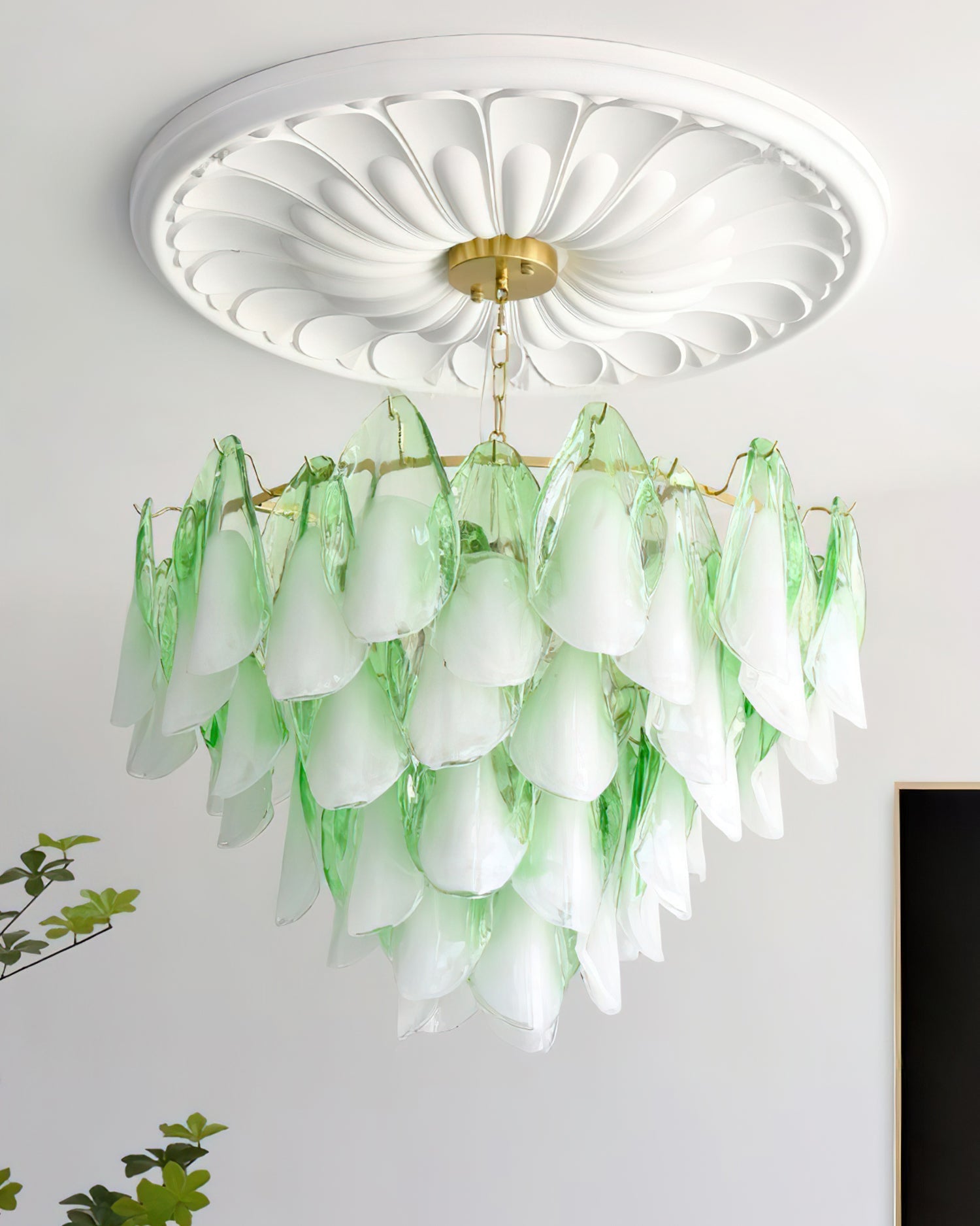 The Clérachic chandelier in sage green glass petals lit in a room corner beside a potted plant.