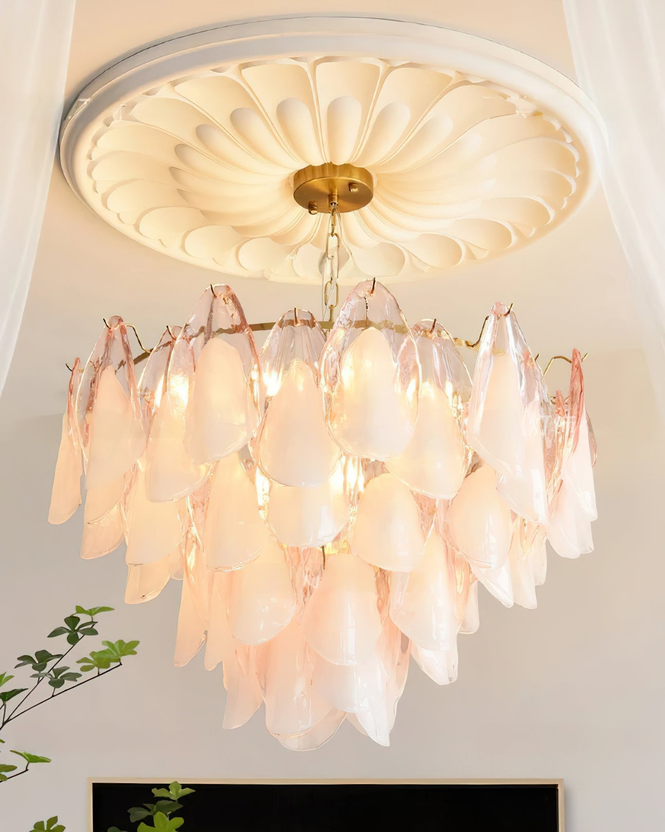The Clérachic chandelier in blush pink and white glass petals lit beside a sheer curtain and plant.