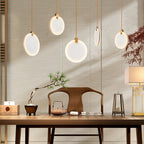 Five lit alabaster disc pendants from Clérachic at varied heights above a wooden desk with a bonsai, a teapot and a table lamp.