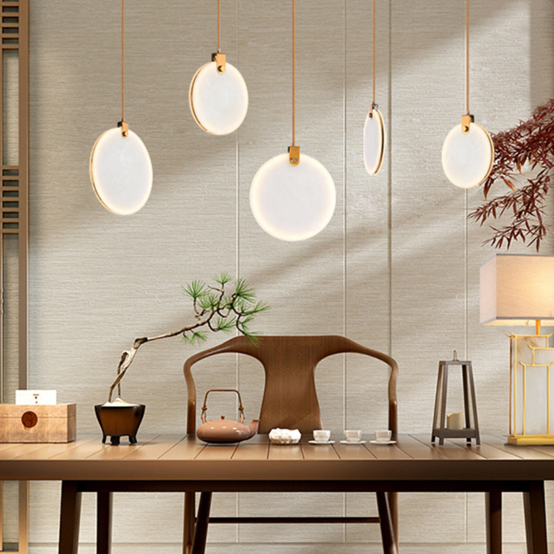 Five lit alabaster disc pendants from Clérachic at varied heights above a wooden desk with a bonsai, a teapot and a table lamp.