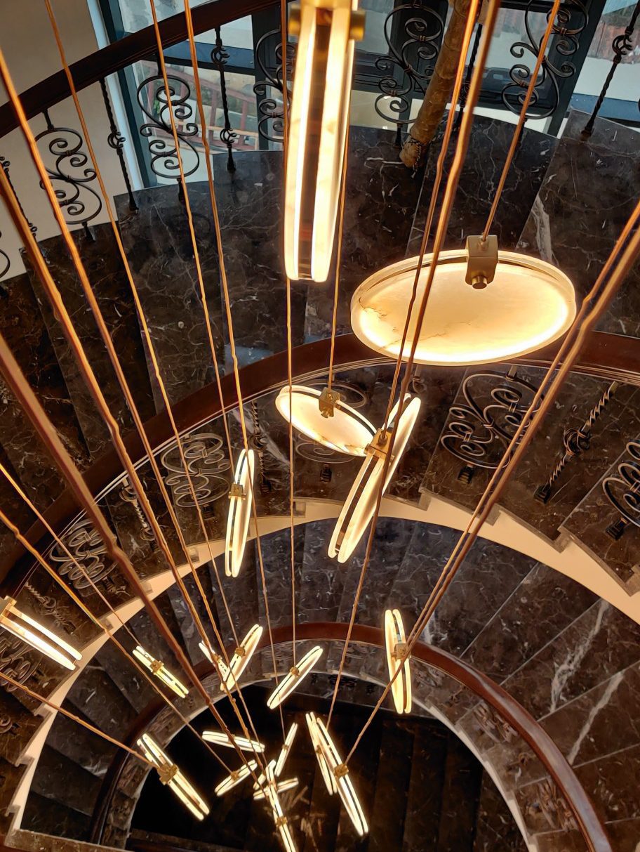 Near the top of a marble spiral staircase, a Clérachic alabaster disc glows beside long tan cords and edge-on discs below.