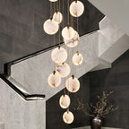 Clérachic alabaster discs of mixed sizes hang over a stair with pale stone walls, a lit handrail and a dark vase of branches.