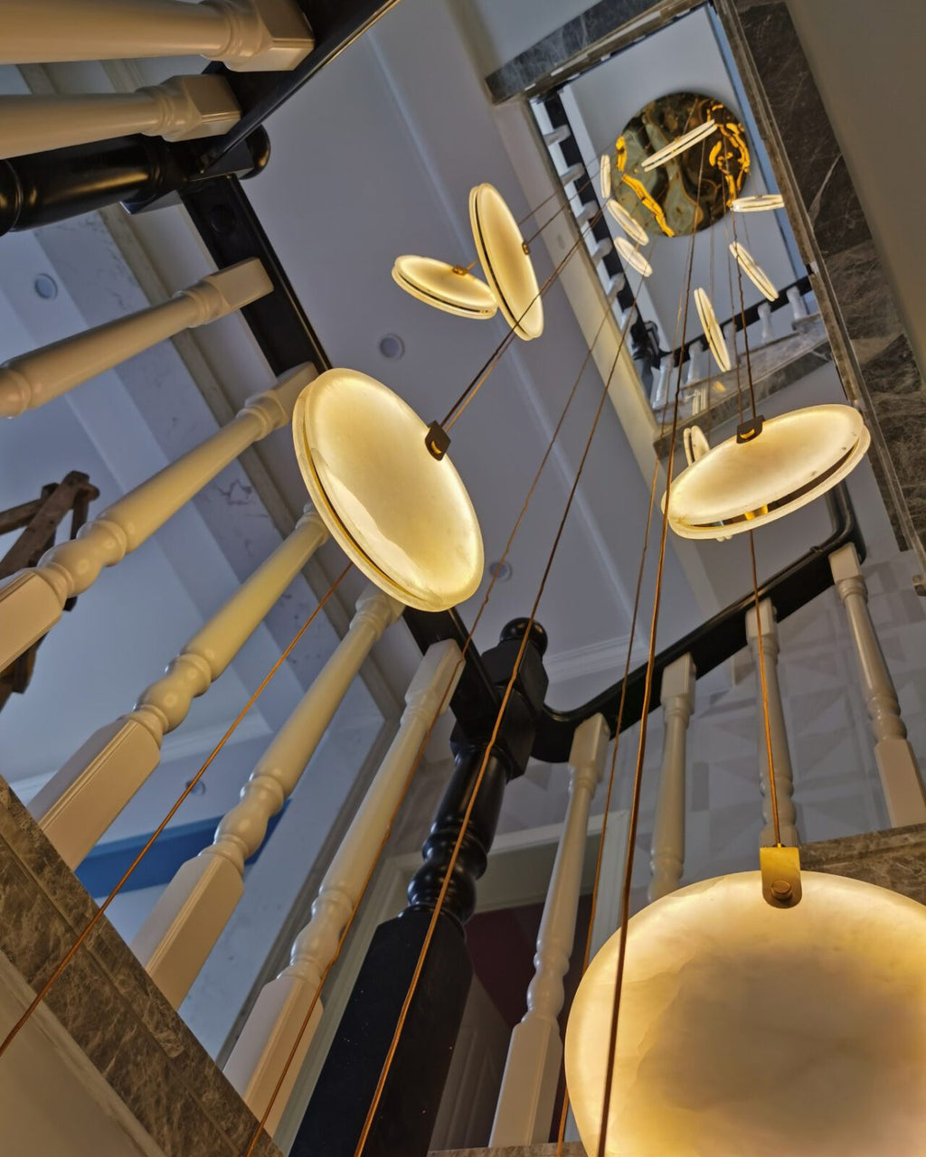 Looking up a stairwell with cream balusters, a Clérachic alabaster disc glows at the bottom of the frame beneath a gold plate.