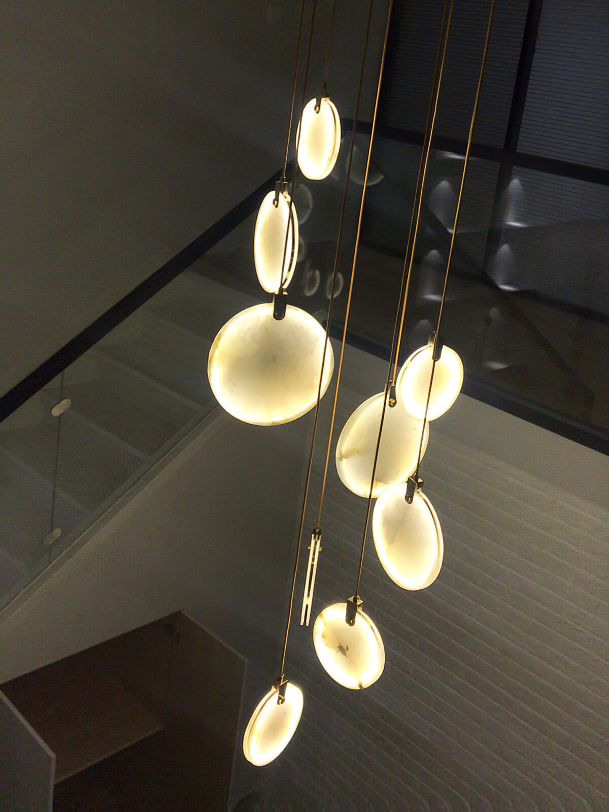 Lit Clérachic alabaster discs on tan cords hang in a dark stairwell, with a glass panel and a pale wall at the left.