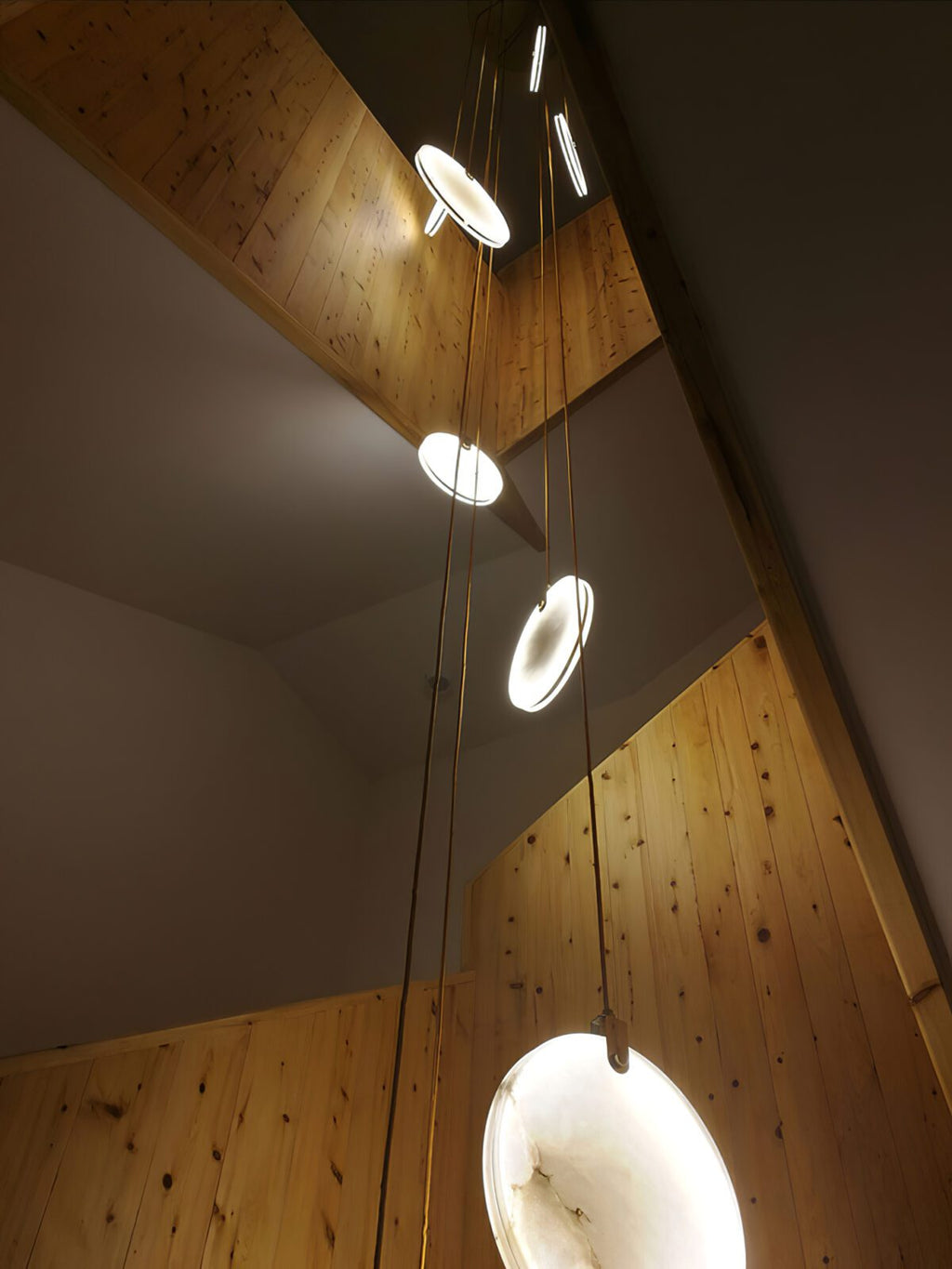 Clérachic alabaster discs glow in a stairwell lined with pale knotty timber, with a bright disc in the lower right.