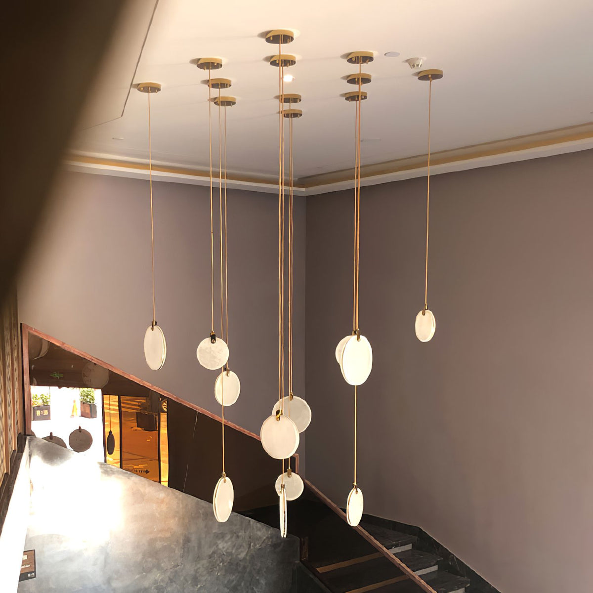 Clérachic alabaster disc pendants hang at varied lengths from several separate gold canopies in a tall stair hall with mauve walls.