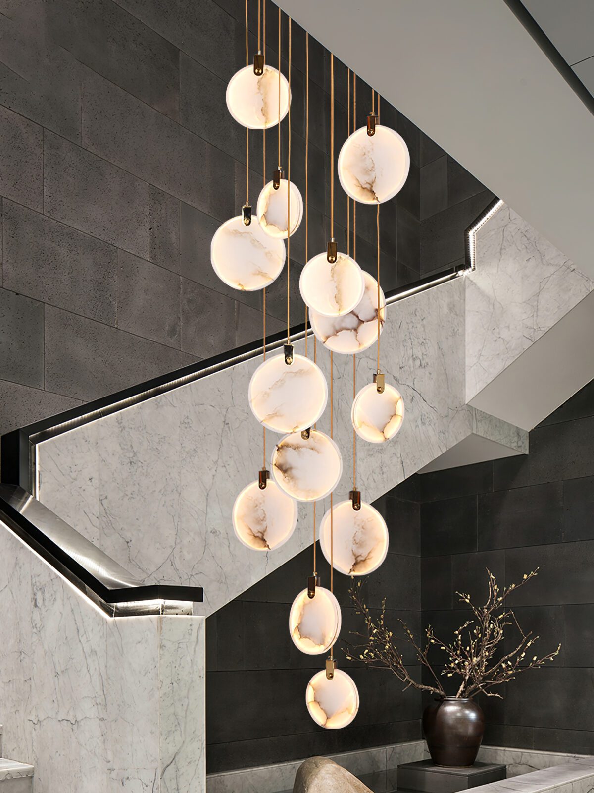 A Clérachic cluster of lit alabaster discs hangs over a stair of pale stone with a dark vase of branches at the foot.
