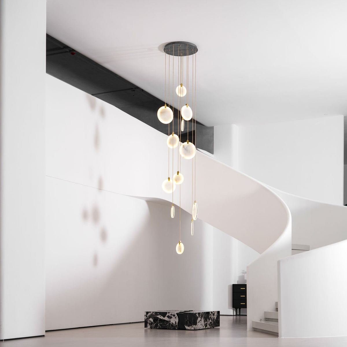 A Clérachic alabaster disc cluster on a dark ceiling plate hangs beside a white sculptural staircase with a black marble block below.