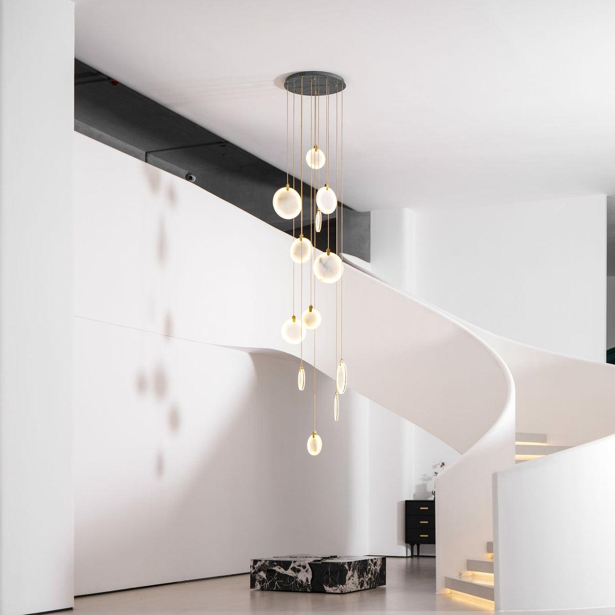 Clérachic alabaster disc pendants of a tall cluster hang in a white hall, with a curved stair and a black marble block on the floor.