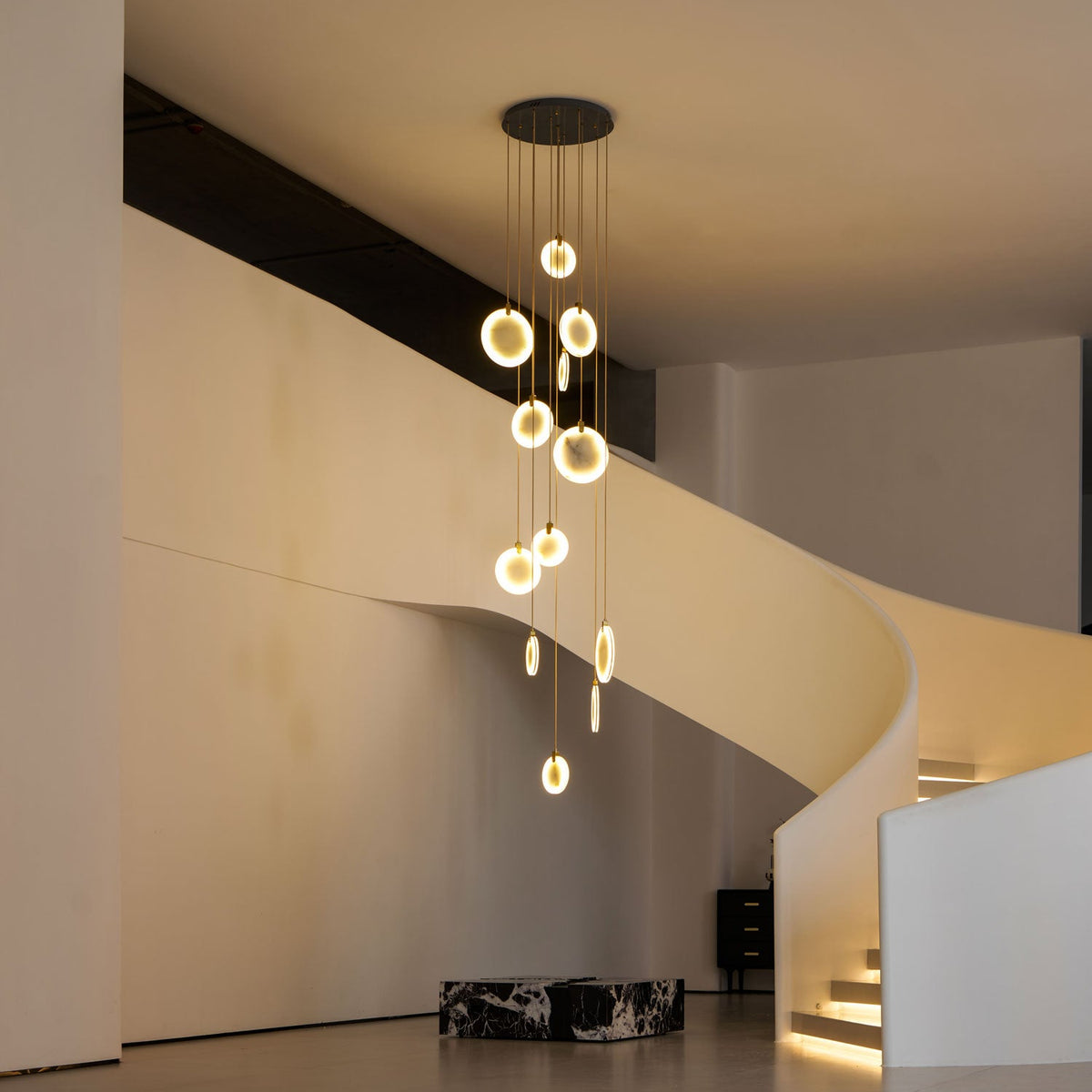 A Clérachic alabaster disc cluster glows warm gold in a white curved-stair hall, with a black ceiling plate and lit steps.