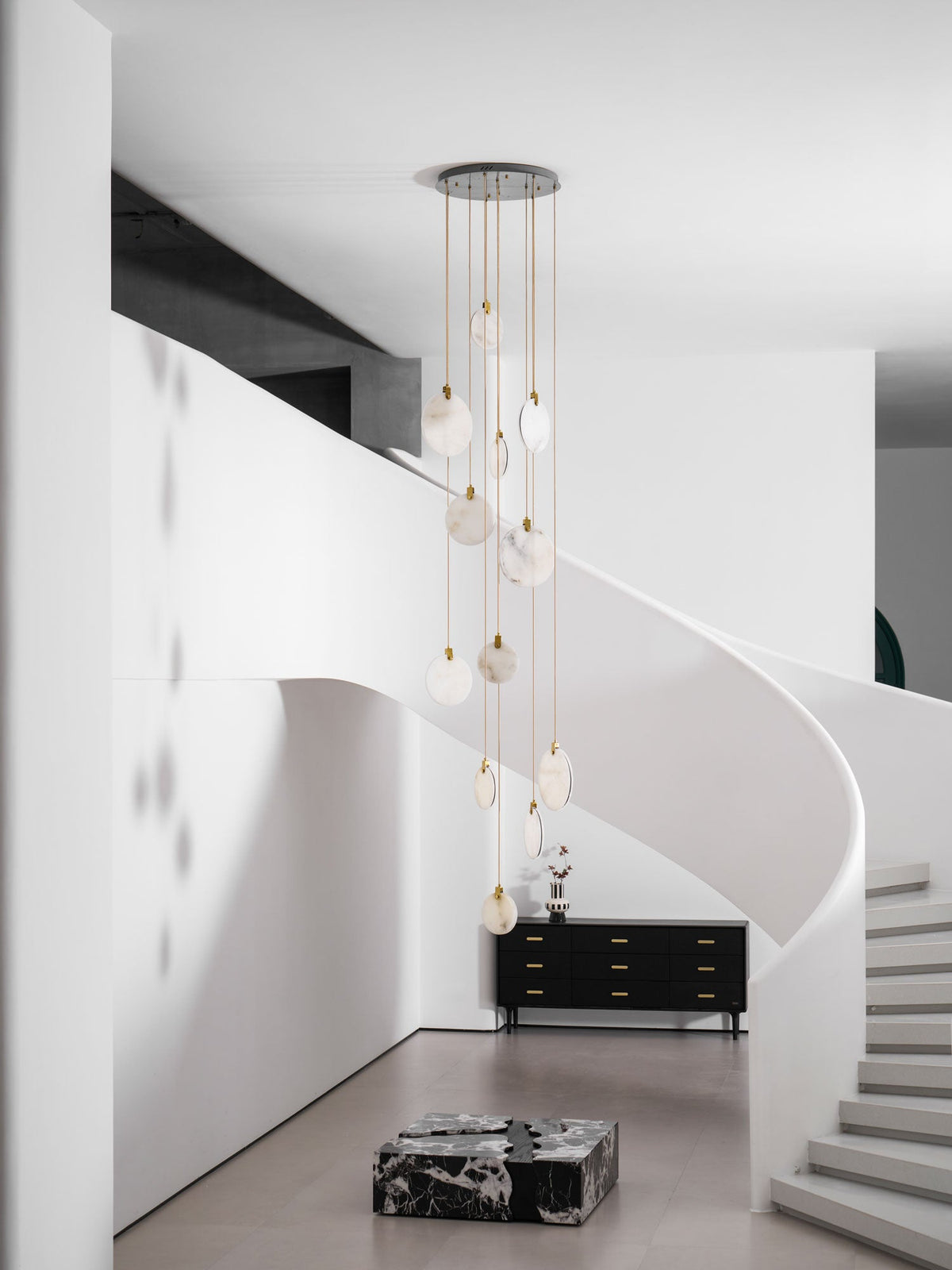 Unlit Clérachic alabaster discs, pale and milky, hang from a dark ceiling plate in a white hall with a curving stair.