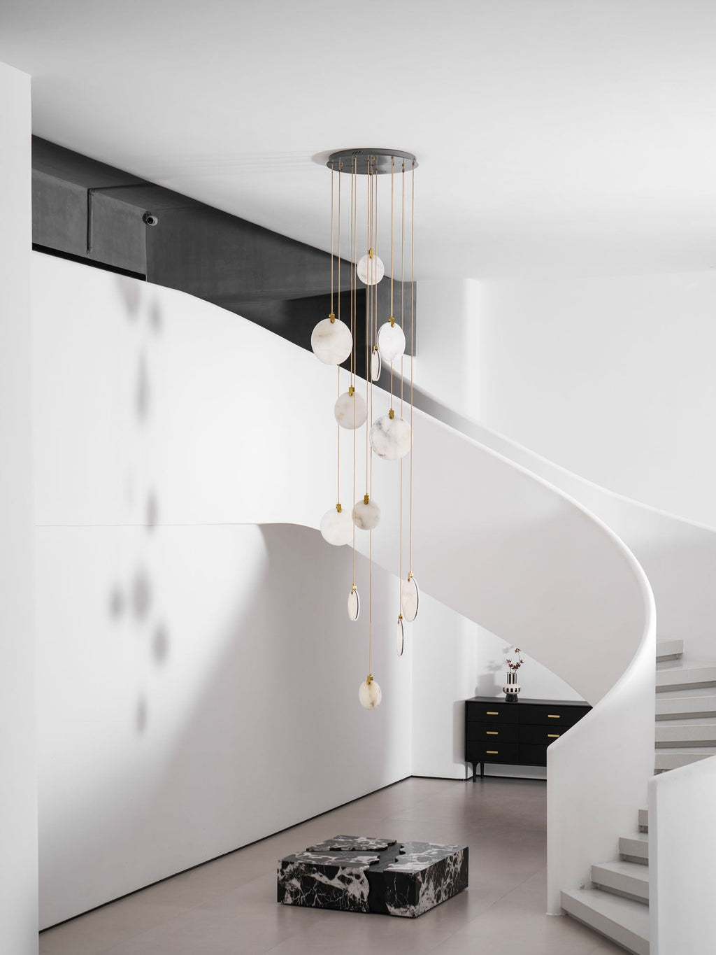 Clérachic alabaster discs unlit in a daylit white stair hall, hanging beside a black sideboard and a marble block.