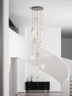A tall Clérachic cluster of unlit alabaster discs on gold cords hangs in front of a white curved stair, above a black sideboard.