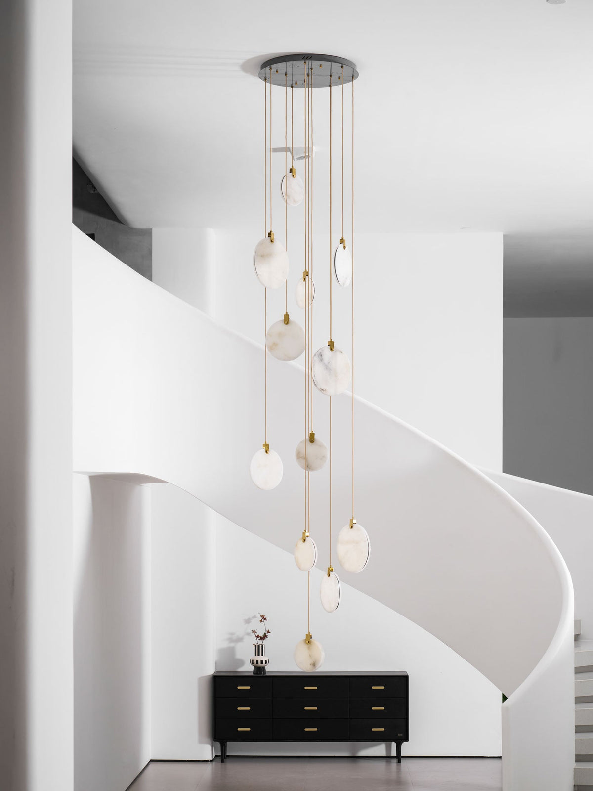 A tall Clérachic cluster of unlit alabaster discs on gold cords hangs in front of a white curved stair, above a black sideboard.