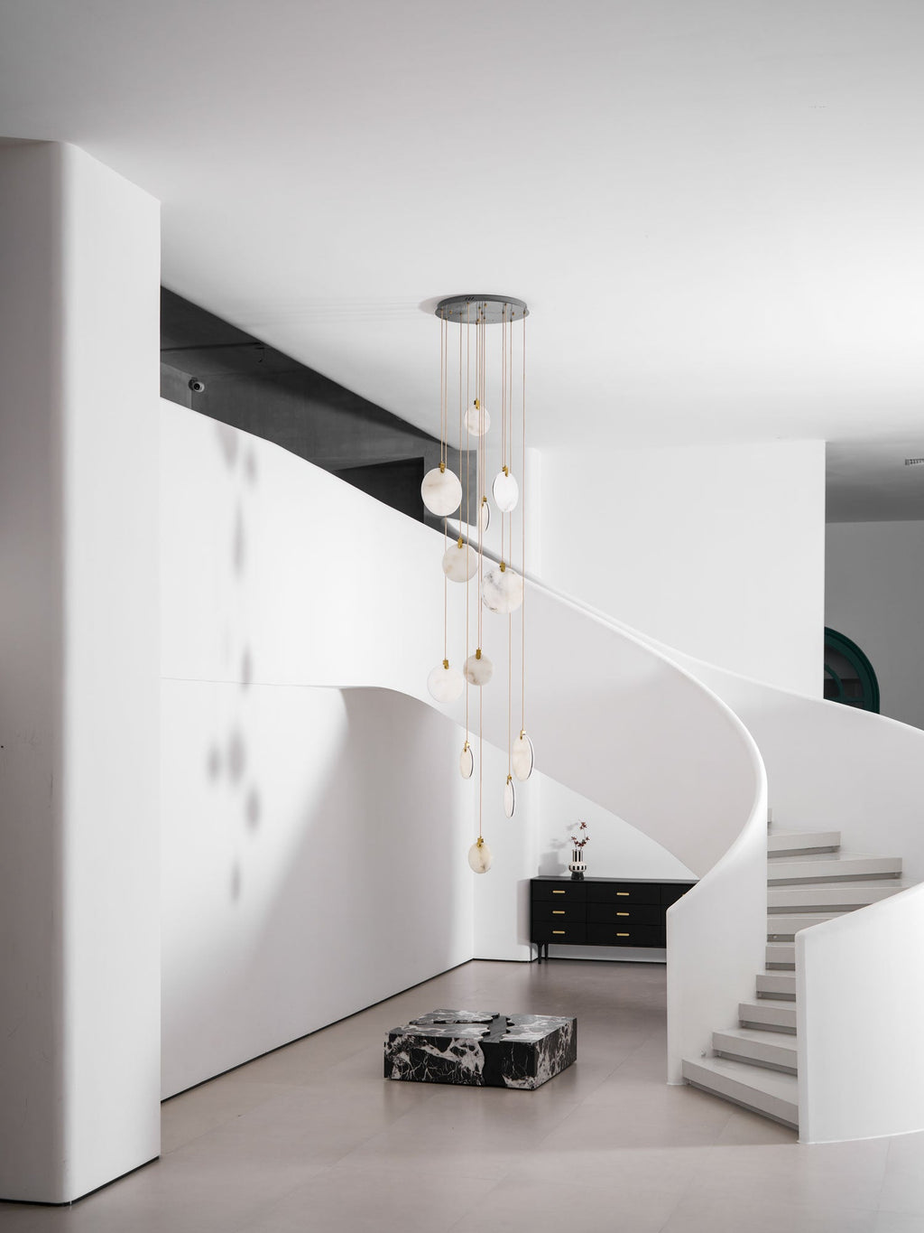 Wide view of an unlit Clérachic alabaster disc cluster in a double-height white room, with a stair at the right and a marble block.