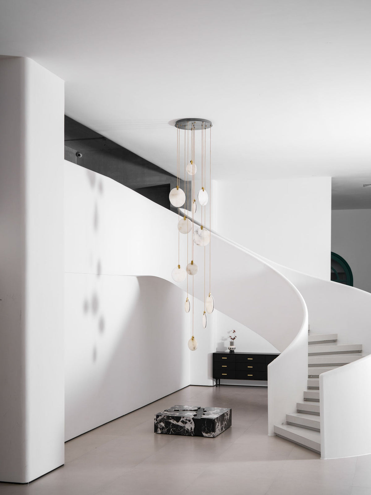 Wide view of an unlit Clérachic alabaster disc cluster in a double-height white room, with a stair at the right and a marble block.
