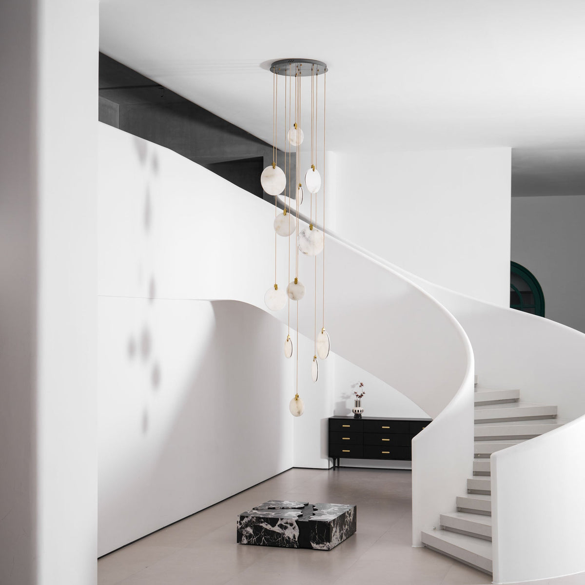 Clérachic alabaster disc pendants hang unlit beside a white curved wall, with a black sideboard and stone-coloured steps.