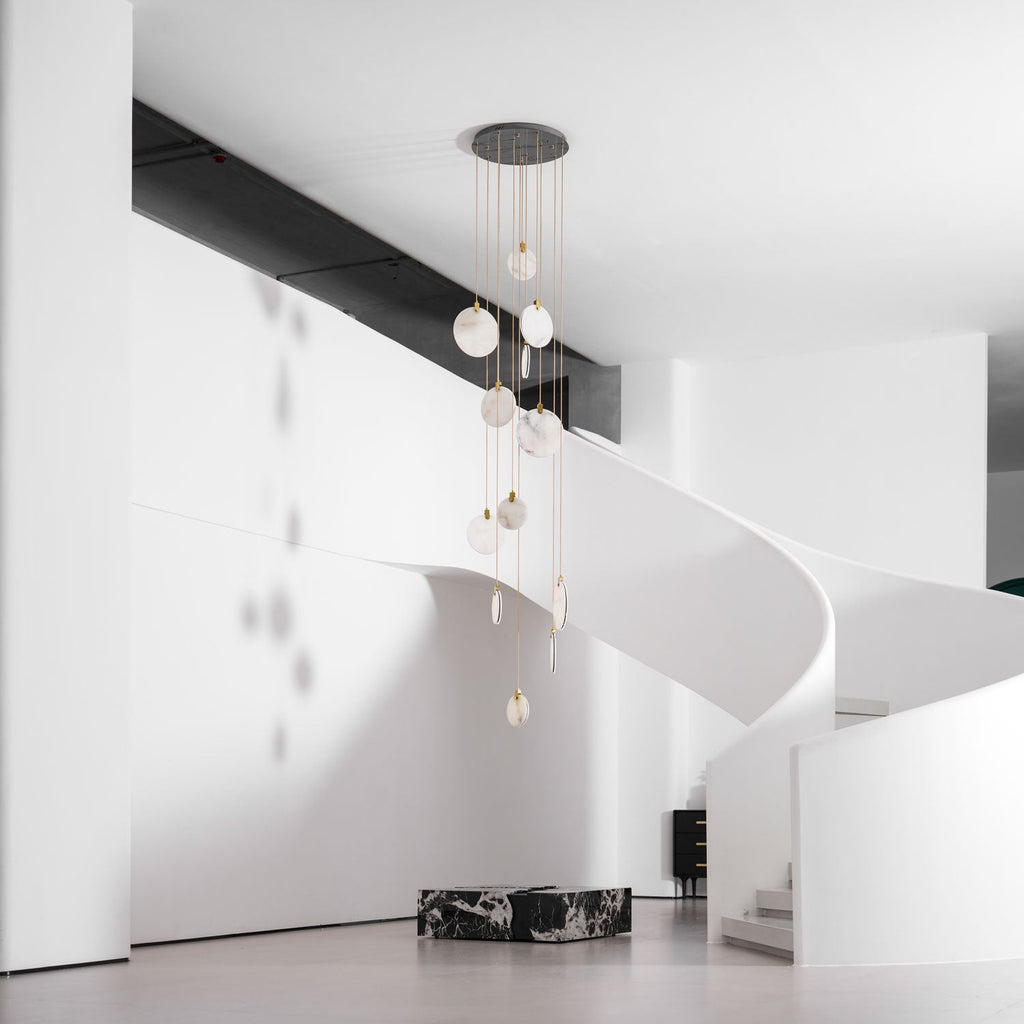 A Clérachic alabaster disc cluster seen unlit from a distance, in a hall with a white curved stair and a dark marble plinth.