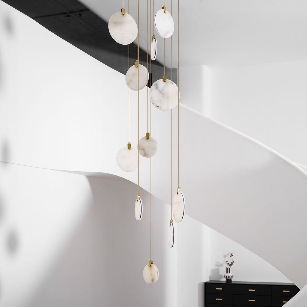 Close view of unlit Clérachic alabaster discs on gold cords in a white stair hall, with a black sideboard in the corner.