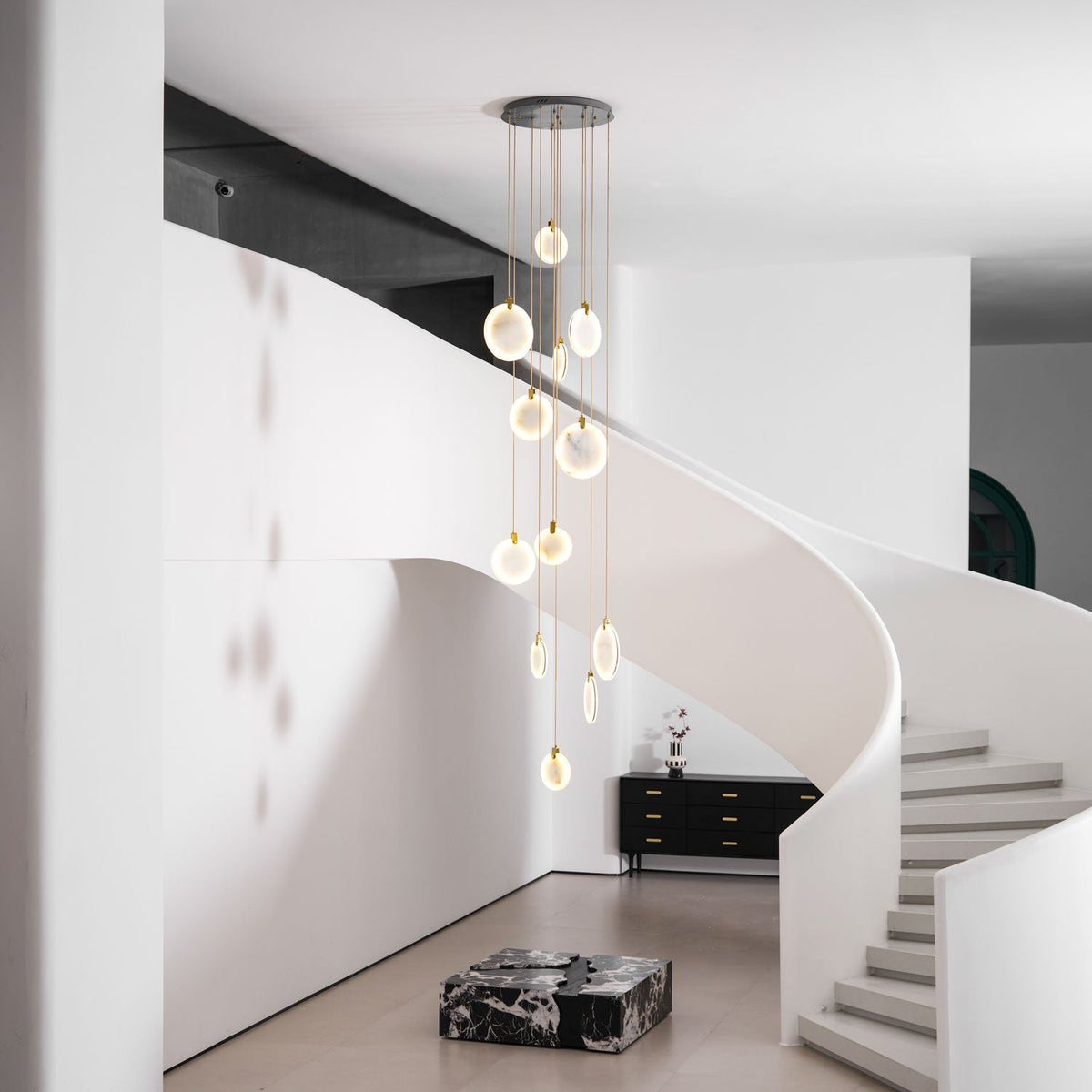 Clérachic alabaster discs lit warm on cords of different lengths, in a white hall with a curved stair and a black sideboard.