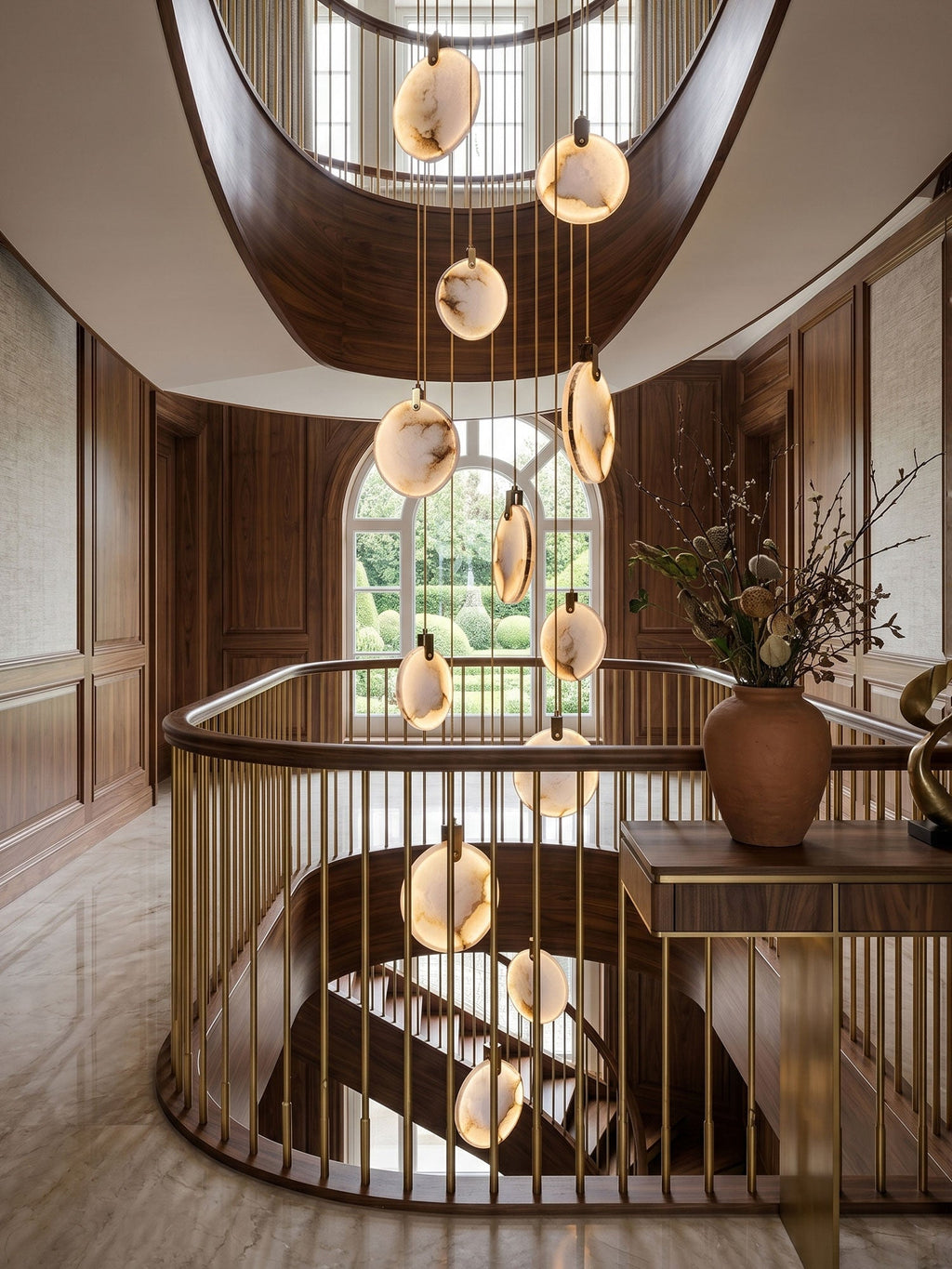 Close view of lit alabaster discs from this Clérachic pendant in a stairwell, with a brass handrail and a large vase of dried stems.