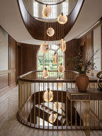 Close view of lit alabaster discs from this Clérachic pendant in a stairwell, with a brass handrail and a large vase of dried stems.