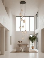 A tall Clérachic cluster of alabaster discs on a dark ceiling plate, in a bright cream double-height room with a plant and two chairs.