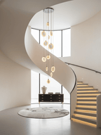 A cluster of alabaster discs from Clérachic hangs inside a spiral cream staircase with lit steps, above a round rug and a black sideboard.