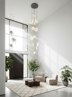 Tall Clérachic alabaster disc cluster on a dark ceiling plate, hanging in a white double-height hall with plants and low chairs.