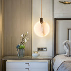 A single Clérachic alabaster disc pendant glows on a cord beside a bed, in front of a timber-panelled wall with a vase of flowers.