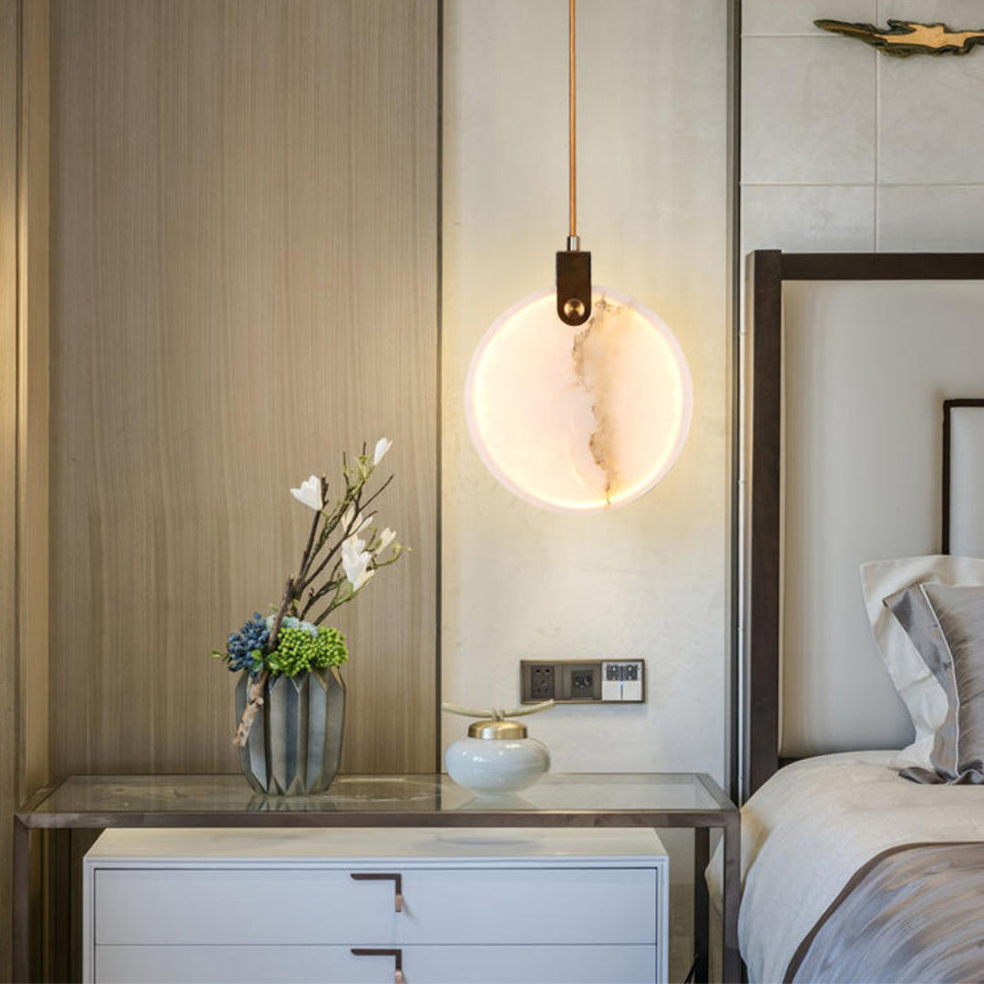 A single Clérachic alabaster disc pendant glows on a cord beside a bed, in front of a timber-panelled wall with a vase of flowers.