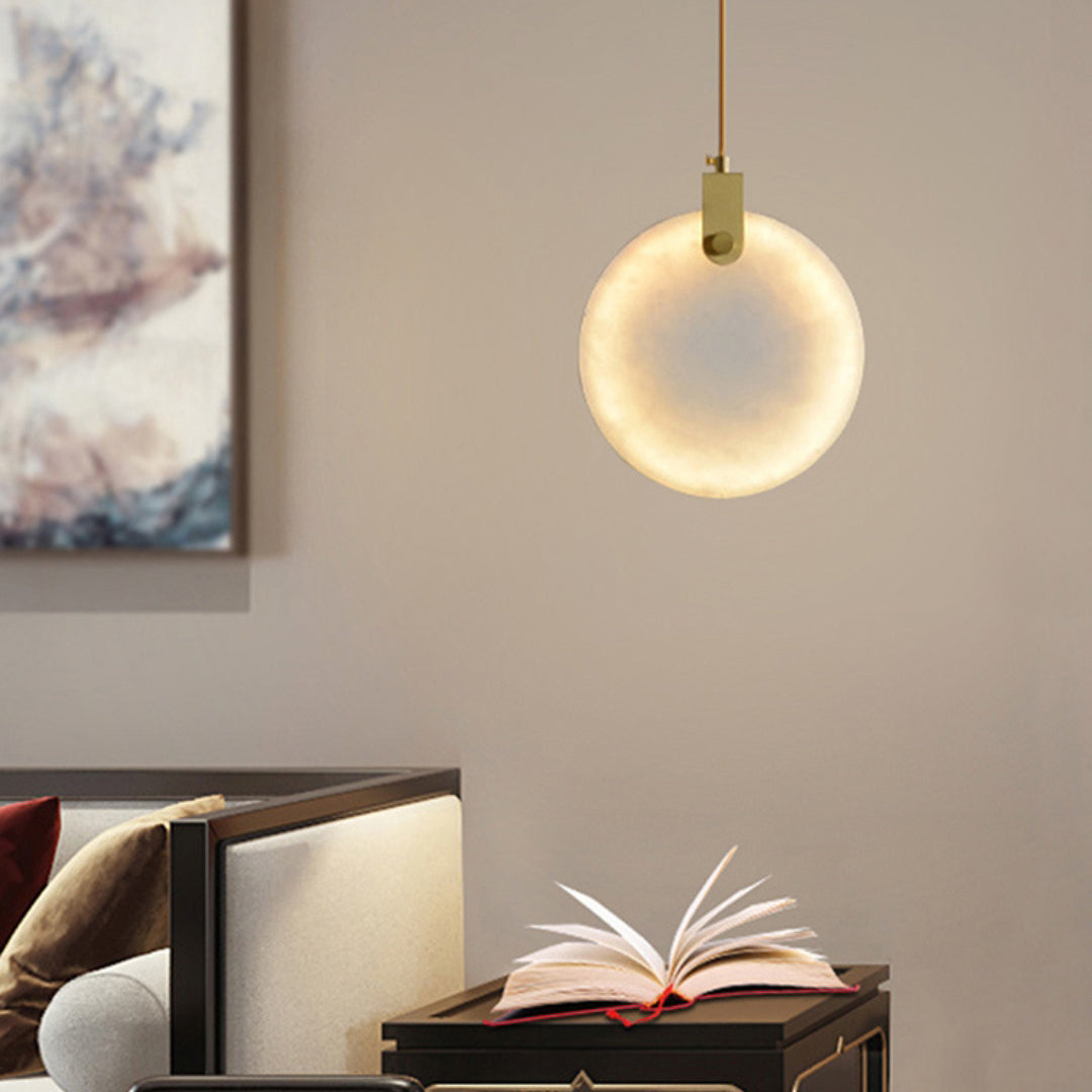One round Clérachic alabaster disc pendant on a brass fitting glows on a taupe wall, above an open book on a dark bedside table.