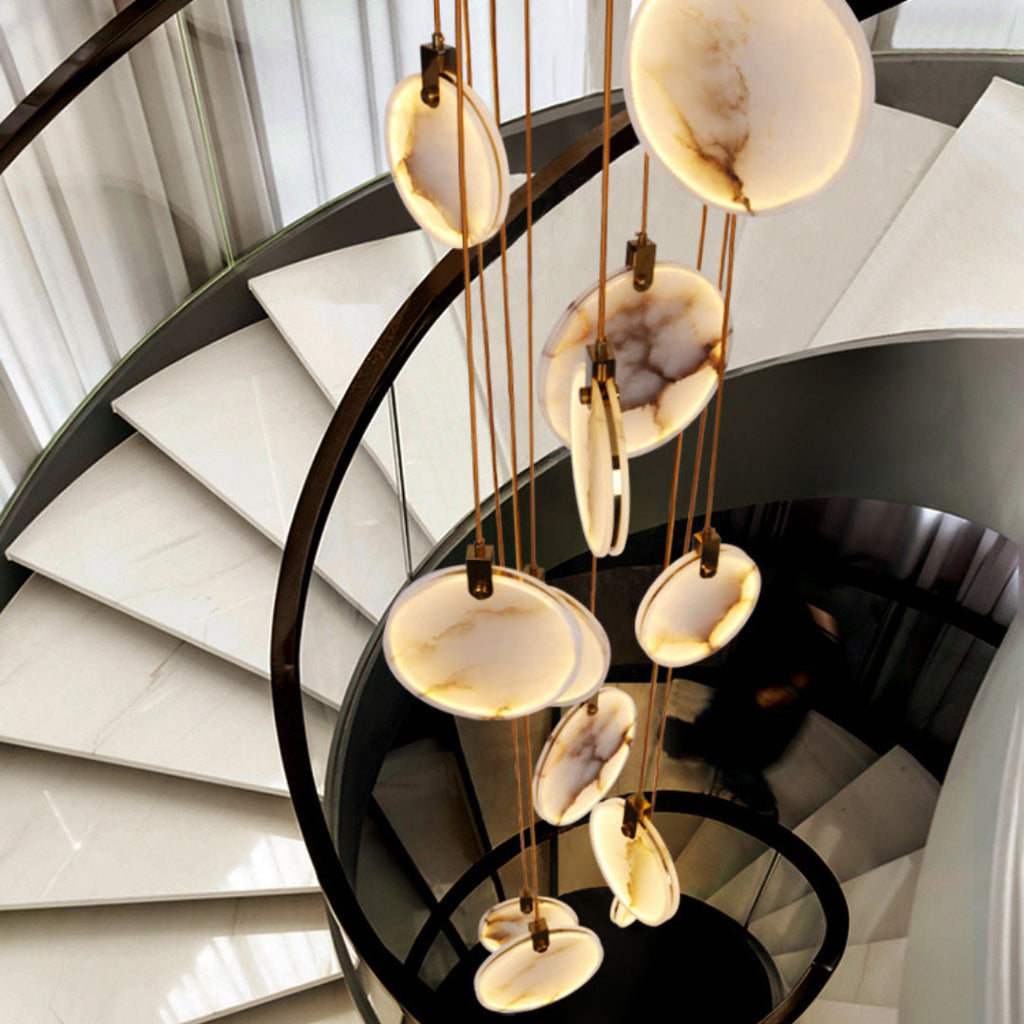 Looking down a spiral staircase of pale stone treads, a Clérachic cluster of alabaster discs hangs on tan cords beside a black rail.