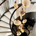 Looking down a spiral staircase of pale stone treads, a Clérachic cluster of alabaster discs hangs on tan cords beside a black rail.