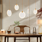 Five lit Clérachic alabaster disc pendants at varied heights over a timber table with a pine bonsai, a teapot and a table lamp.