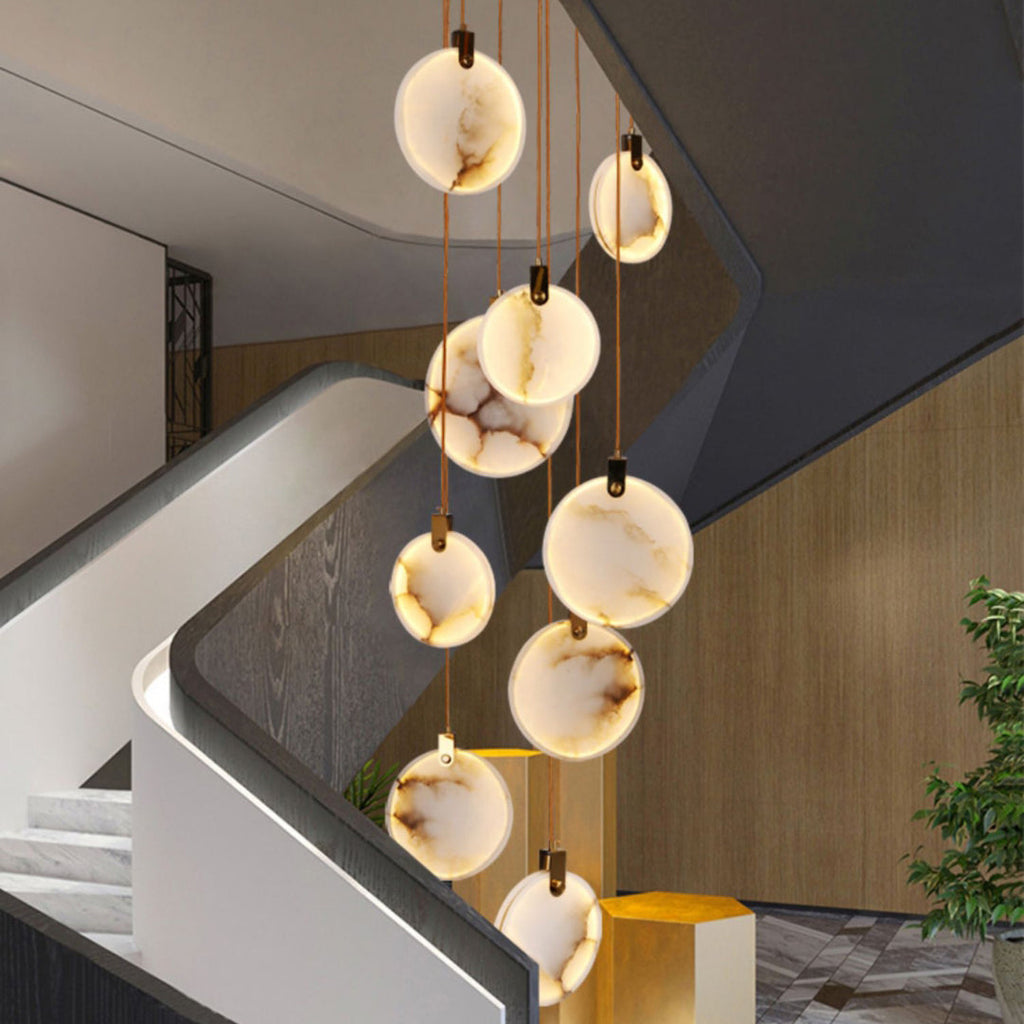 Veined alabaster discs of this Clérachic pendant hang beside a white curving stair and a timber-clad wall, with a plant at the right.