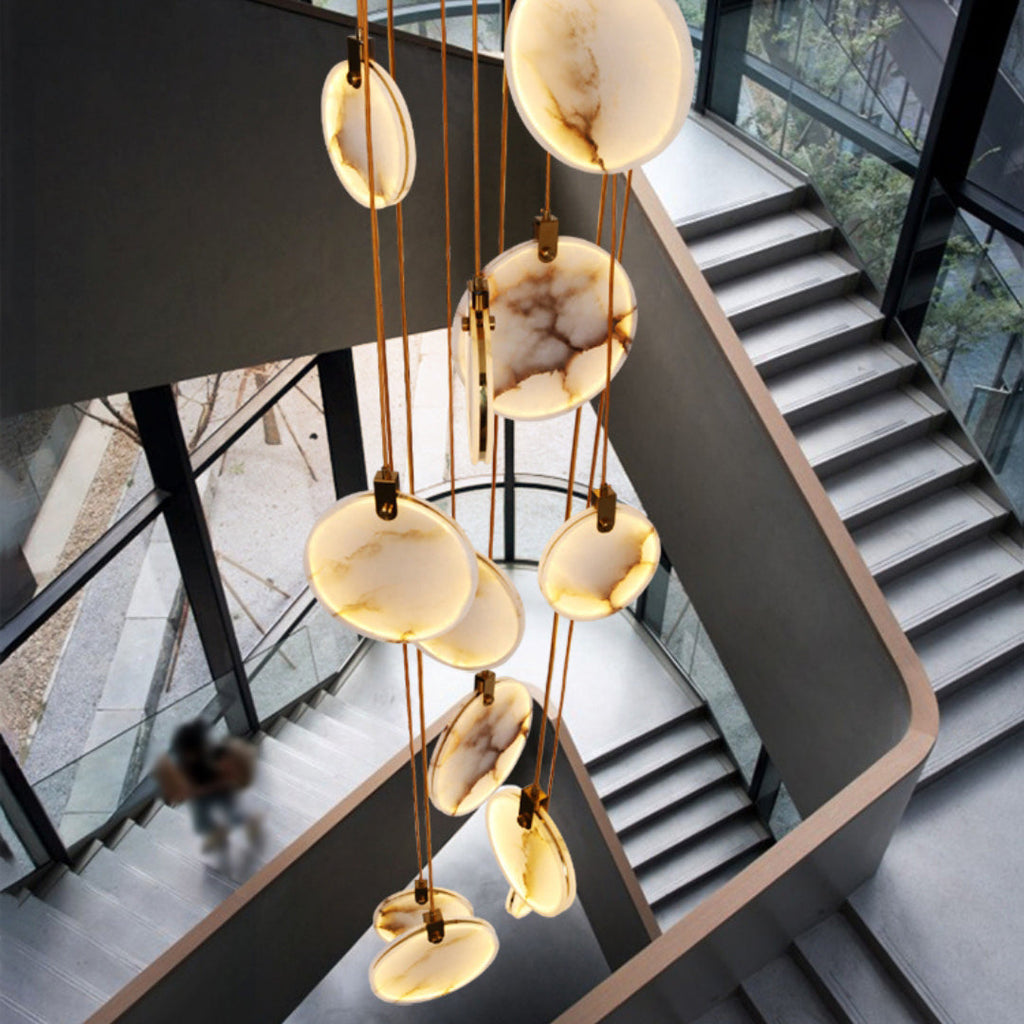 Seen from above, Clérachic alabaster disc pendants hang through a stairwell with dark steel stairs and a glass wall, with a blurred figure below.