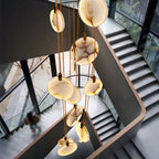 Seen from above, Clérachic alabaster disc pendants hang through a stairwell with dark steel stairs and a glass wall, with a blurred figure below.