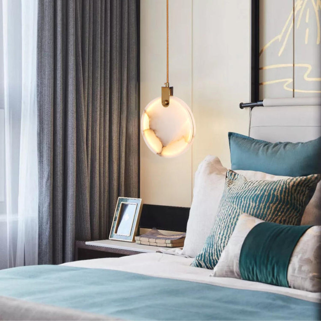 A single Clérachic alabaster disc pendant glows above a bed with teal and cream cushions, beside a framed photo on the bedside table.